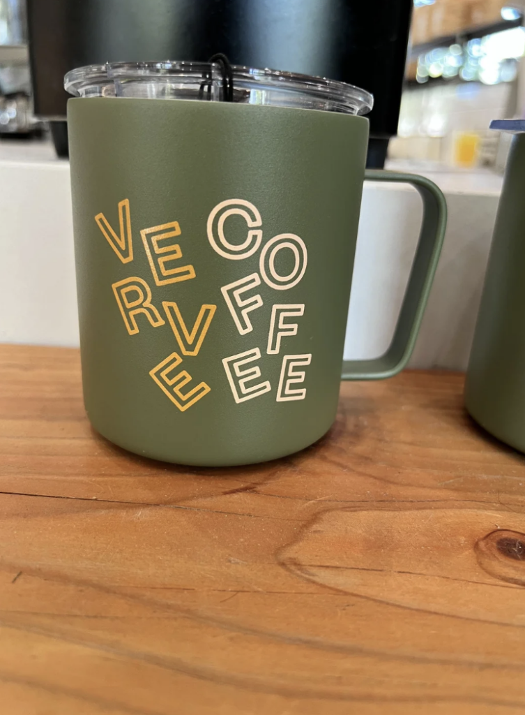A green insulated coffee mug with a clear lid rests on a wooden surface. The mug features the word "COFFEE" in a playful, scattered arrangement with yellow and white letters. The background shows a blurred café setting.