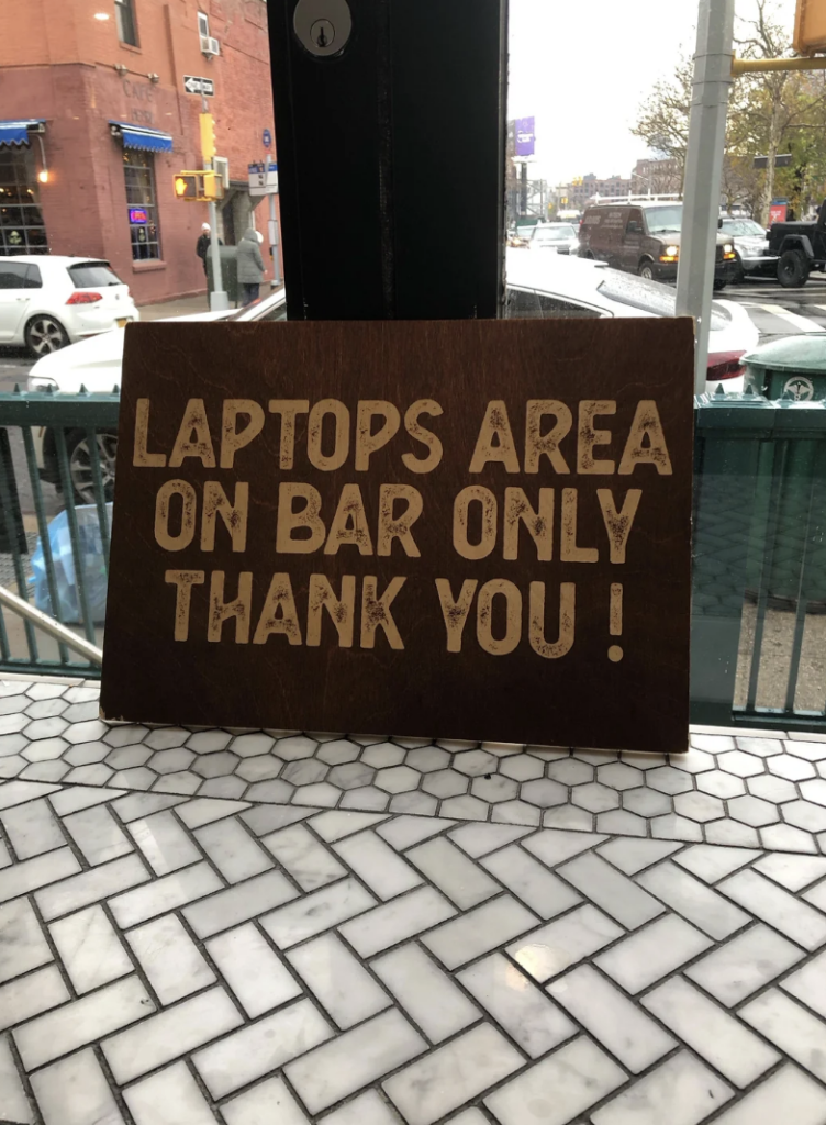 A wooden sign on a white herringbone-tiled table reads, “Laptops area on bar only thank you!” In the background, outdoor seating and a street with cars and buildings are visible.
