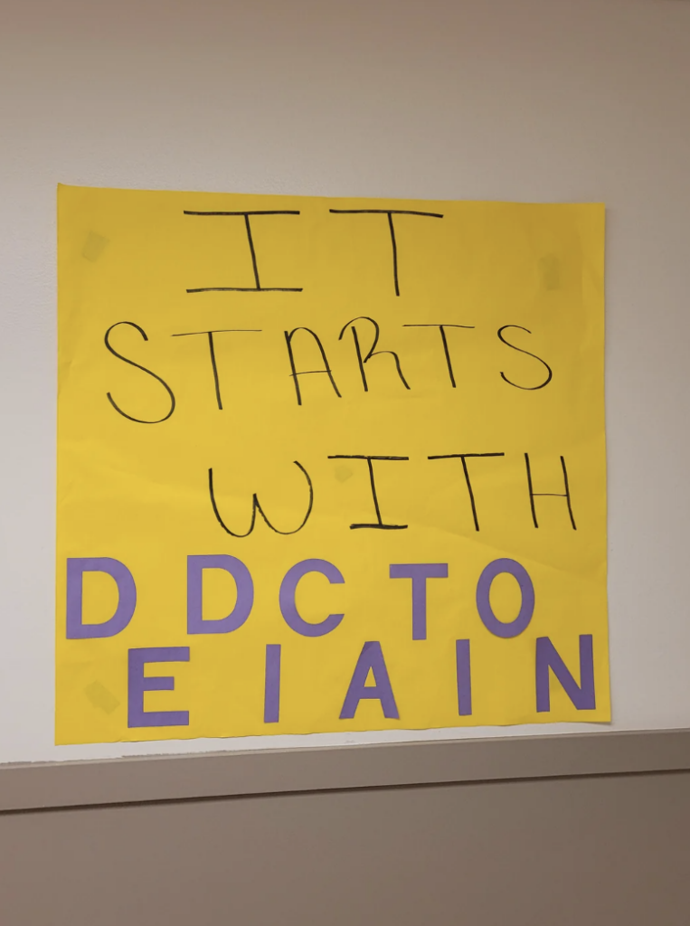 A yellow poster with the phrase "IT STARTS WITH" written in black marker at the top. Beneath that, there are scrambled letters "DDCTO EEIAIN" in purple. The poster is taped on a cream-colored wall above a ledge.