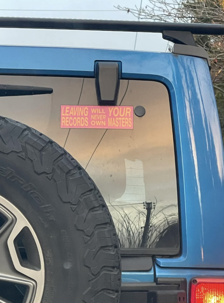 The back of a blue vehicle featuring a bumper sticker on the rear window that reads, "Leaving your records will never own masters." Part of a spare tire is visible on the left, and a utility pole can be seen in the reflection on the window.