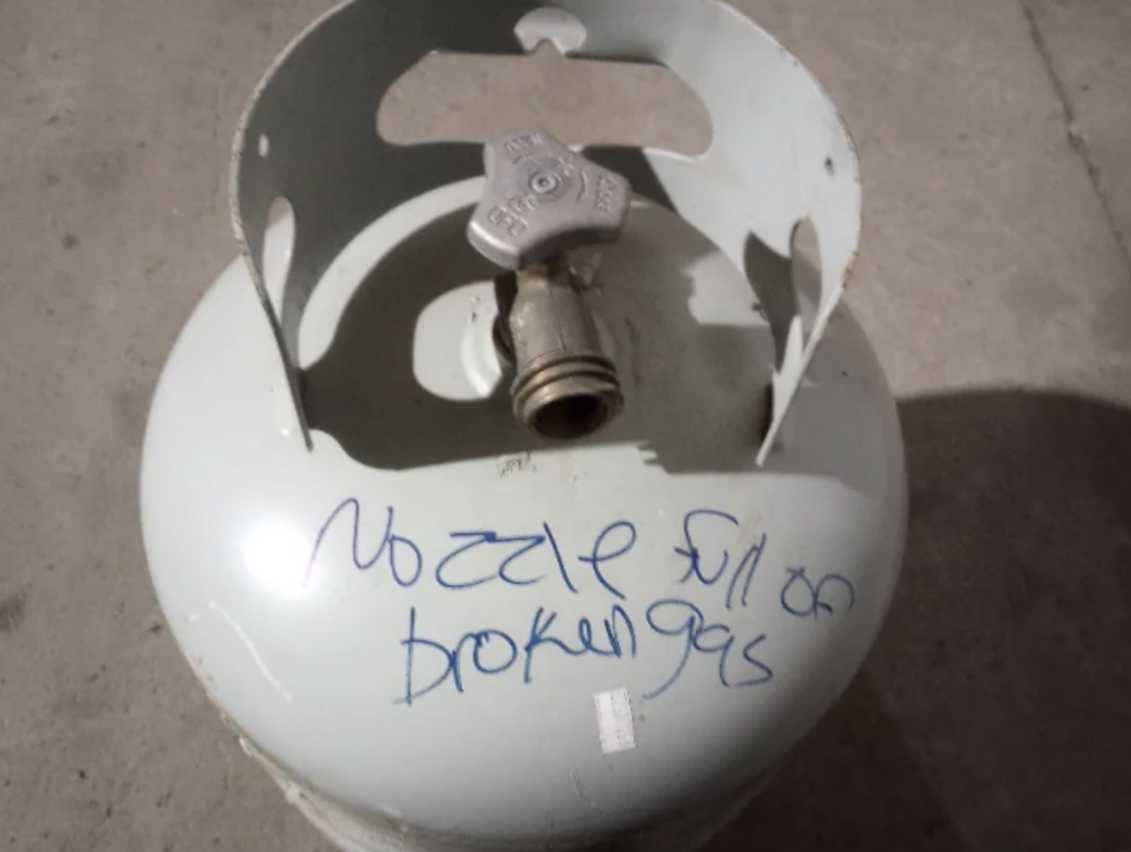 A white gas cylinder with a metal valve on top, placed on a concrete surface. The words "Nozzle full or broken gas" are handwritten on the cylinder in blue marker. The top guard ring of the cylinder has some discoloration and scratches.