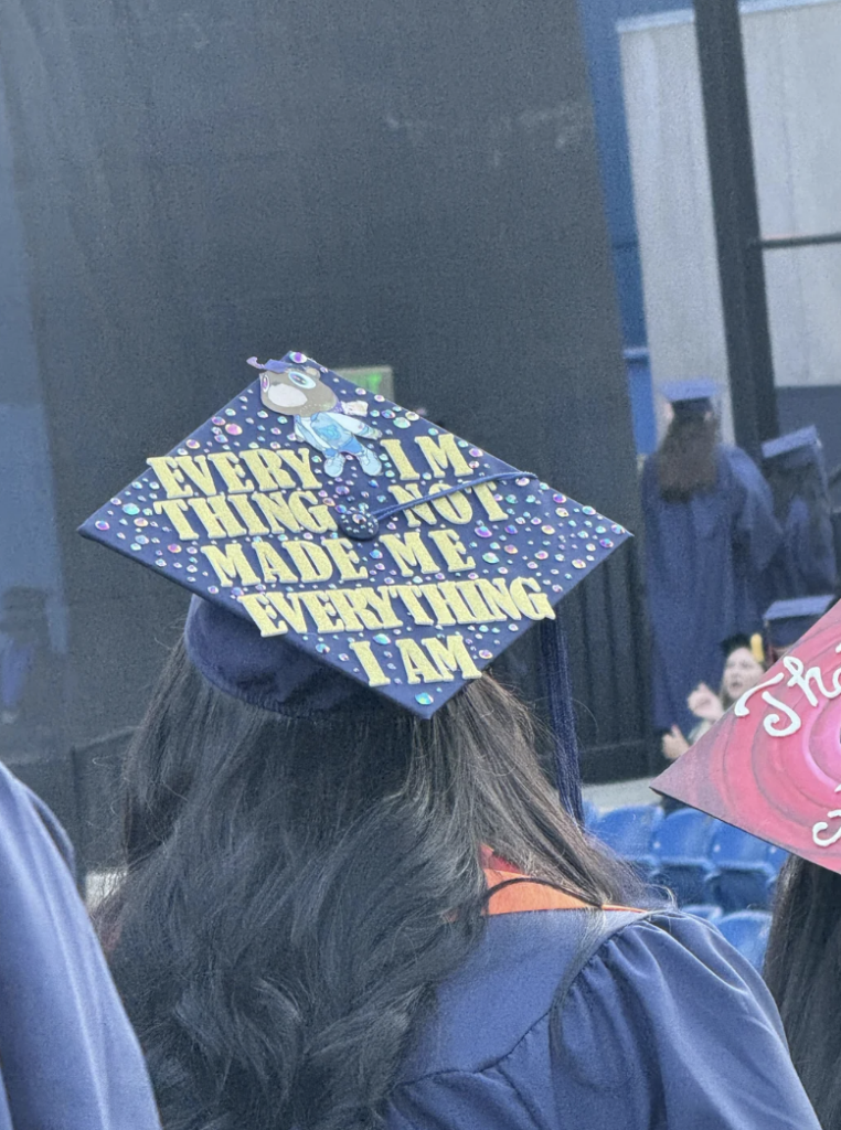 A graduate wears a decorated cap with the words "Everything I’m thinking about made me everything I am" written in yellow. The cap features a small character and colorful decorations. The graduate is surrounded by other graduates wearing blue gowns.