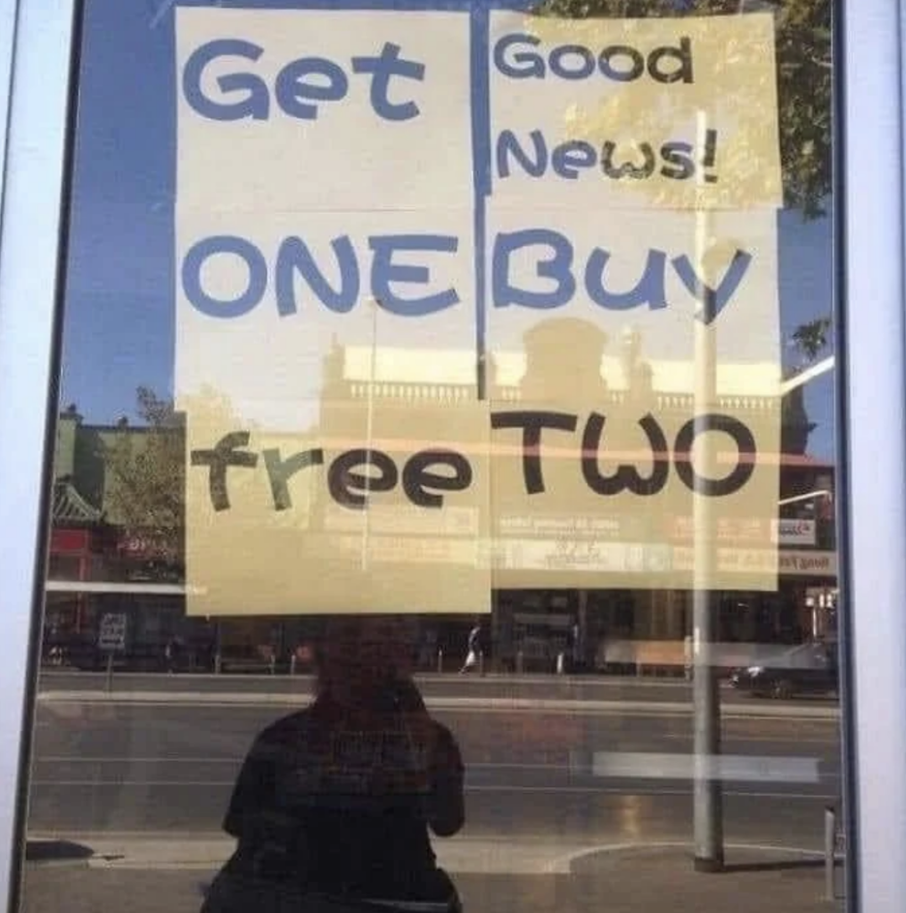 A store window shows a confusing promotional sign that reads, "Get Good News! ONE Buy free TWO." The reflection of the street and buildings outside can be seen in the window.