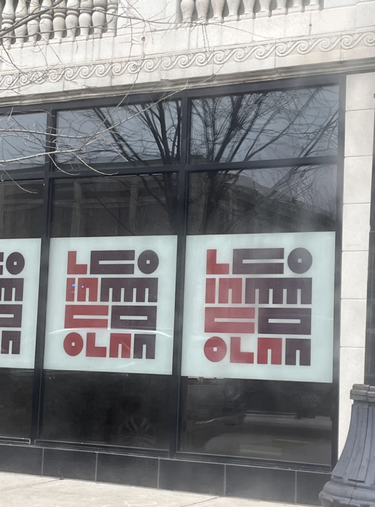 Storefront windows with large, blocky letters spelling out "Up Urban Salon" in a minimalist, abstract style. The letters are stacked and colored in shades of red and brown. A tree is reflected in the glass, and architectural details are visible at the top.