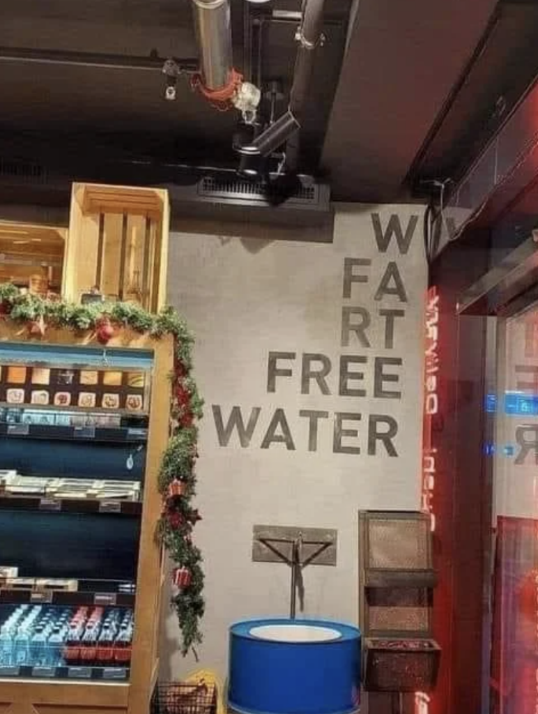 A store display shows a sign on the wall that humorously spells “WATER” vertically with the phrases “WATER,” “FAT FREE,” and “FREE WATER” formed with overlapping letters. Nearby shelves hold various products, and a blue display container sits in the foreground.