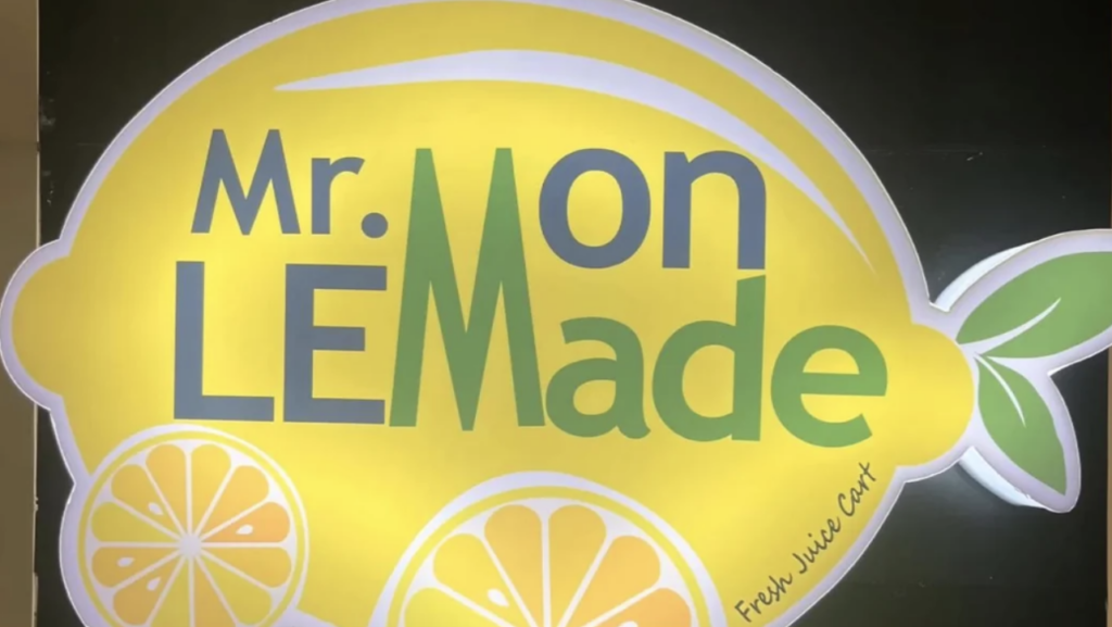 Image of a sign shaped like a lemon with the text "Mr. Lemon LE Made" in green and yellow. There are lemon slice illustrations at the bottom, and a small leaf on the right. The text "Fresh Juice Cart" is in smaller green text at the bottom right.