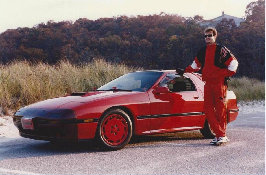 A person in a red and black athletic outfit stands next to a red sports car parked by a roadside with grass and trees in the background. The person rests one hand on the car and looks at the camera, wearing sunglasses and sneakers. The setting appears to be a rural area.