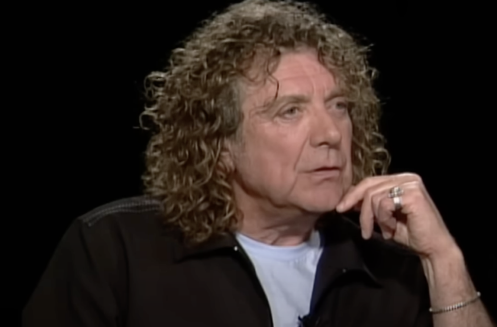 A man with long, curly hair is seated, wearing a black jacket over a white t-shirt. He rests his chin on his hand, looking thoughtful. The background is dark, highlighting his face and upper body. The image conveys a pensive, contemplative mood.