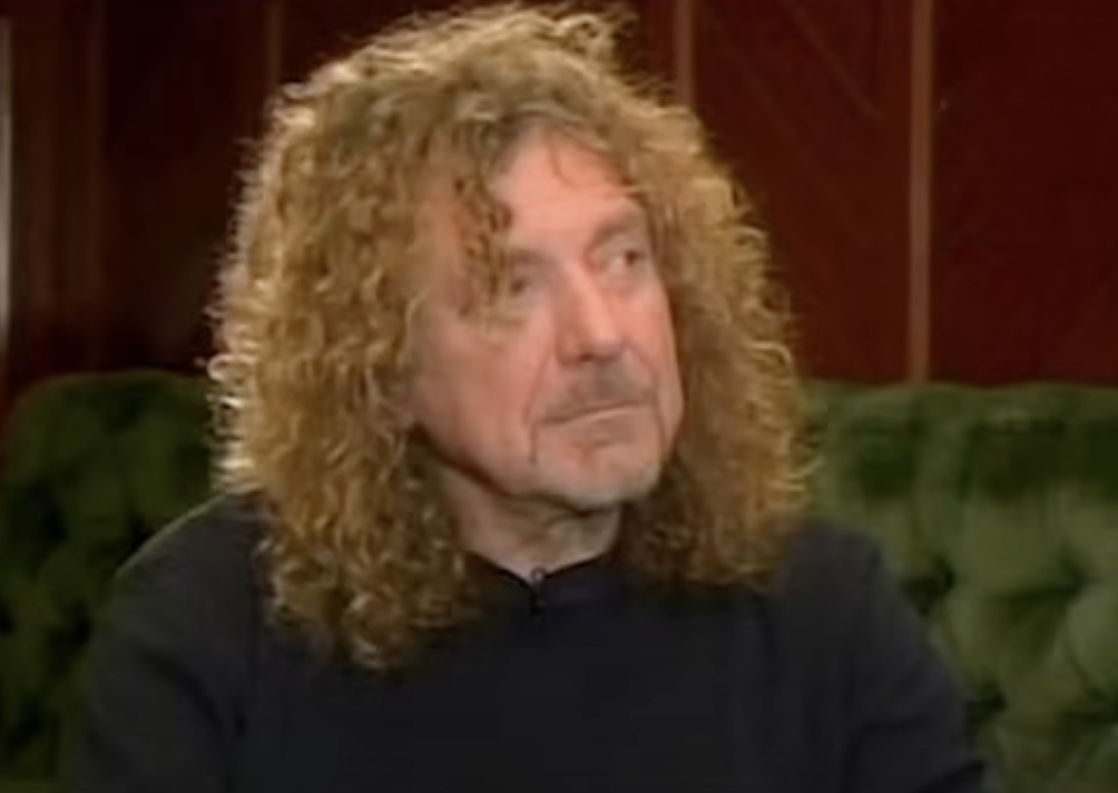 A person with curly, long hair and light facial hair, wearing a dark long-sleeved shirt, is sitting on a green tufted sofa. The background features wooden paneling. They have a thoughtful expression and are looking slightly to the side.