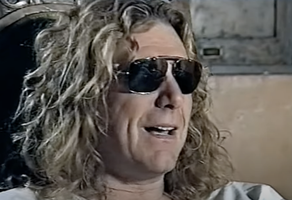 A person with long, curly blond hair is wearing dark sunglasses and smiling. They are seated in an indoor setting, and the background is slightly blurred, featuring what appears to be a wooden structure and a cabinet.