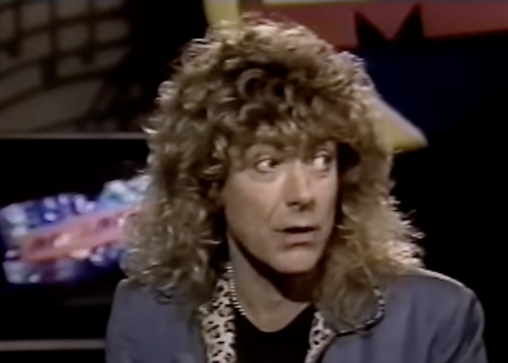 A person with curly, long hair and wearing a blue jacket over a dark shirt appears to be mid-conversation during an interview on a television show. The background features colorful abstract graphics on a screen.