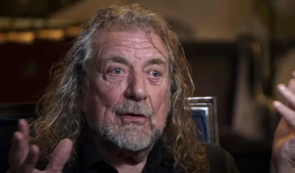 A man with long wavy hair and a beard is seated, gesturing with both hands raised. He has a focused expression and is wearing a dark-colored shirt. The background is slightly blurred, suggesting an indoor setting.