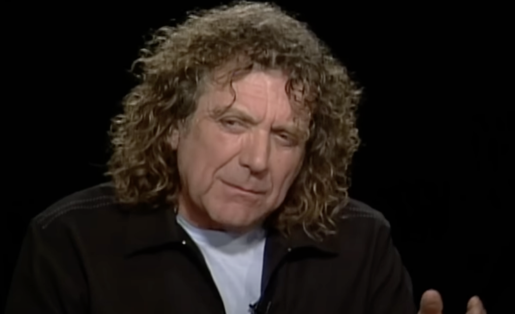 A person with curly, shoulder-length hair and wearing a dark jacket over a light shirt is speaking. The background is black.