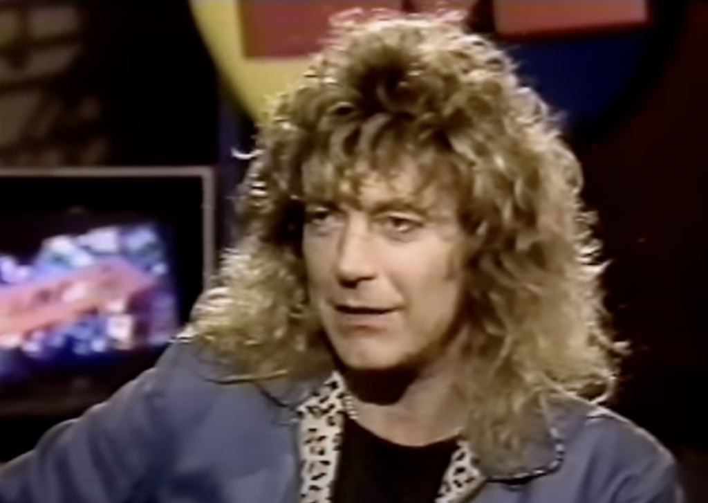 A person with long curly hair and wearing a blue jacket with a patterned collar is seated. The background is dark with a blurry television screen visible behind them. The scene is indoors.