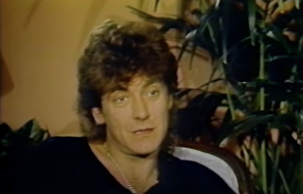 A person with curly hair wearing a black shirt is sitting in front of a background with plants. The person appears to be talking, with a neutral expression.