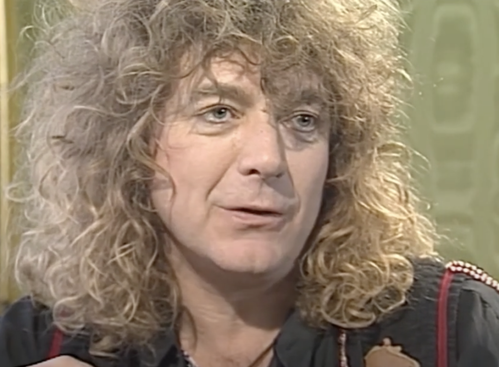 A person with long, curly, blond hair is speaking. They are wearing a dark top and are captured in a close-up shot, which reveals their expressive face against a muted, patterned background.