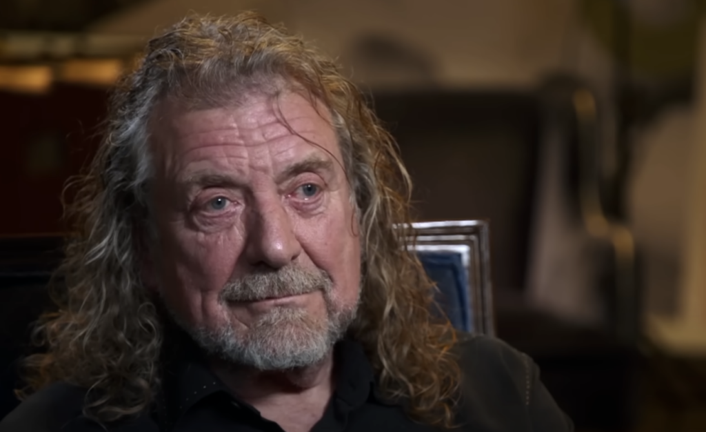 A senior man with long, wavy, light brown hair and a beard sits in a room. He wears a dark shirt and looks slightly to his left, with a thoughtful expression. The background is dimly lit and out of focus, featuring various furniture and decor.