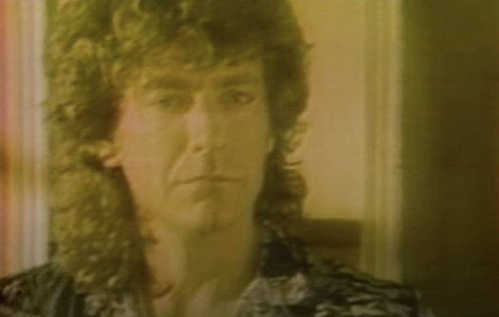 A person with curly, long hair is looking directly at the camera in this image. The image has a warm, yellowish tint and appears slightly grainy, giving it a vintage feel. The individual is wearing a dark patterned shirt.