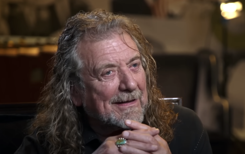 A man with long, wavy gray hair and a beard is sitting with his hands clasped under his chin. He is smiling and looking slightly to the side. He is wearing a dark-colored shirt and a turquoise ring on his right hand. The background is blurred.