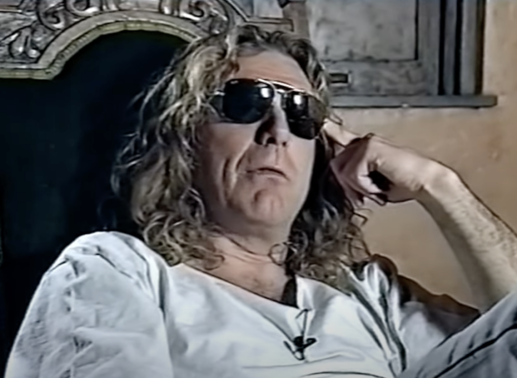 A person with long curly hair is sitting on a chair, wearing dark sunglasses and a light-colored shirt. They appear to be speaking or giving an interview in a vintage setting with a stone wall and arched wooden window behind them.