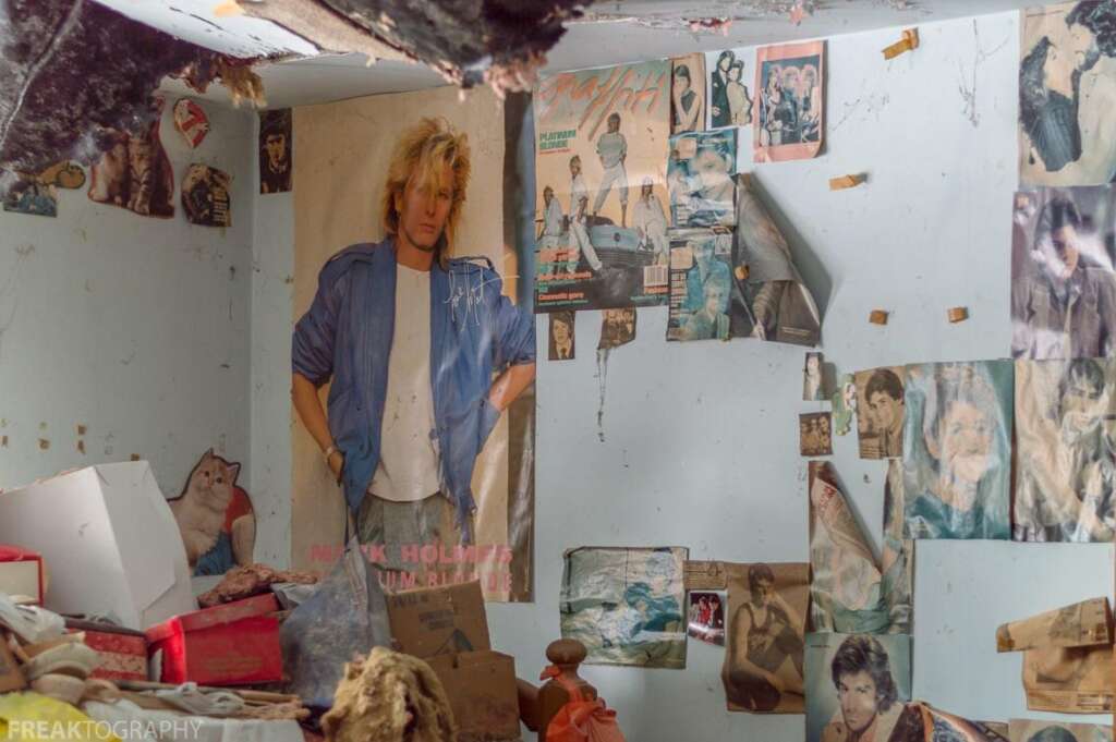 A cluttered room with walls covered in posters of celebrities and a cat. The ceiling is damaged and shows signs of severe water damage. Scattered items and debris cover the floor and an old dresser.