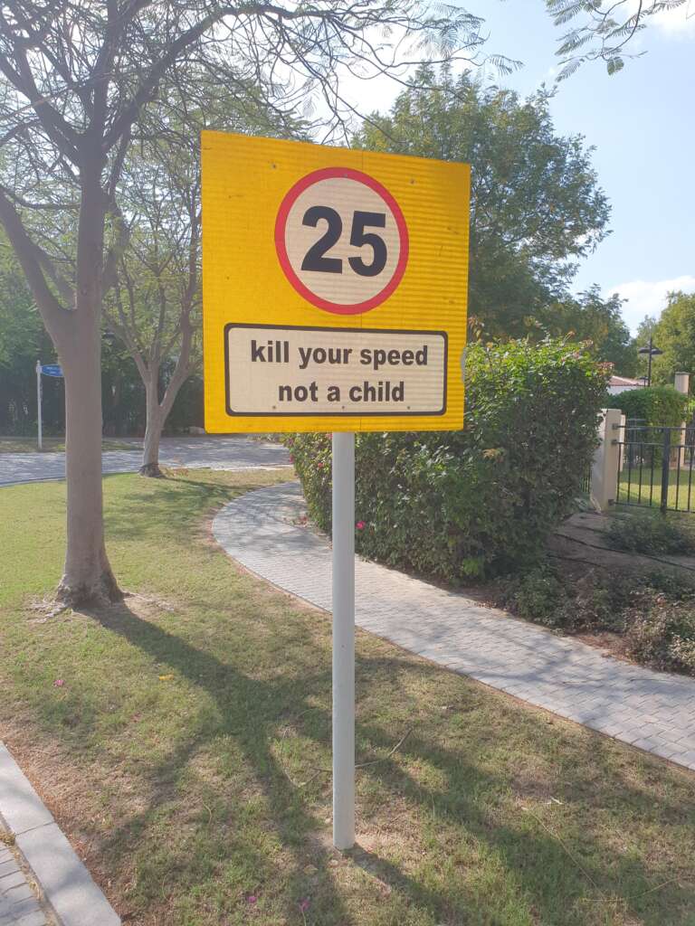 A clear yellow road sign with a red-bordered circle showing the speed limit of 25 mph. Below, a cautionary message reads, "Kill your speed, not a child." The background features a park-like setting with trees, grass, and a sidewalk.