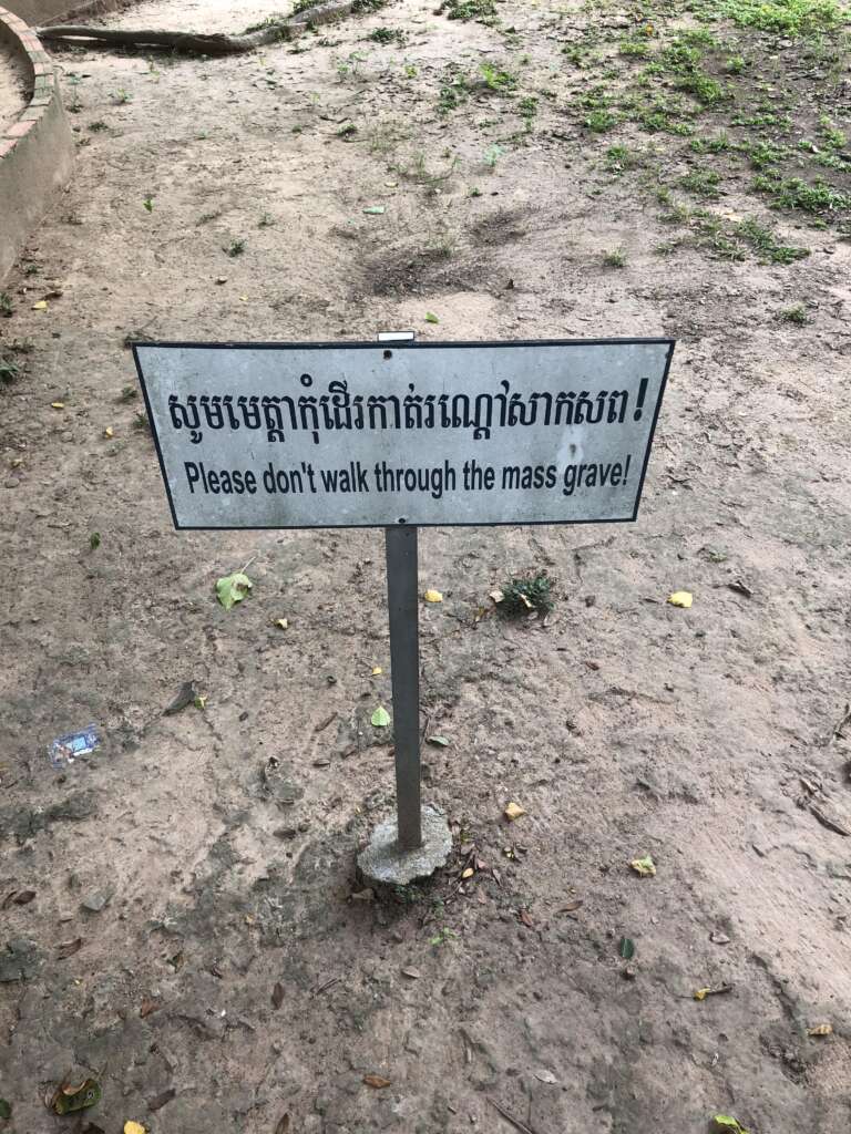 A standing metal sign with a white background warning, "Please don't walk through the mass grave!" is placed on a dirt ground with some patches of small green plants. The text is written in both Khmer and English.