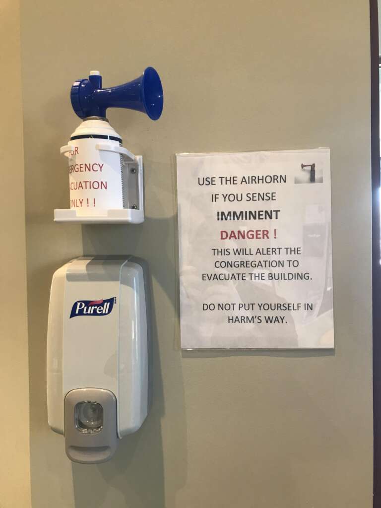 A wall-mounted air horn with a sign that reads: "USE THE AIRHORN IF YOU SENSE IMMINENT DANGER! This will alert the congregation to evacuate the building. Do not put yourself in harm's way." Below it, a Purell hand sanitizer dispenser is attached to the wall.