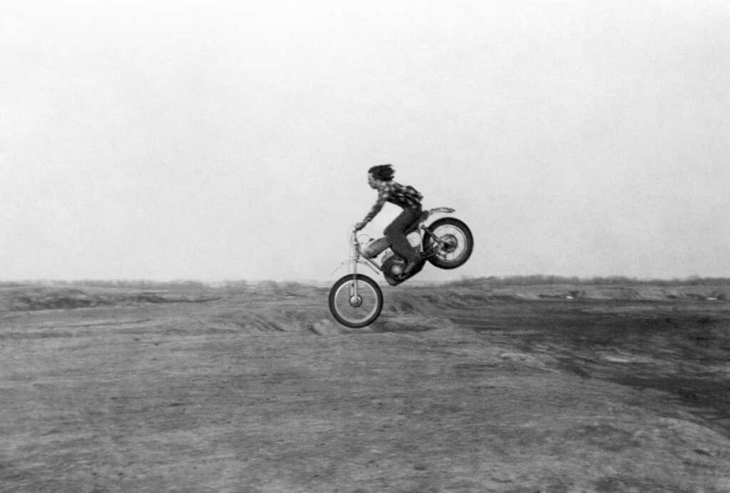 Black and white image of a person in mid-air performing a motorcycle jump. The rider wears a checkered shirt and jeans, riding a dirt bike on a rugged terrain with a clear sky background. The scene captures the high-flying action and thrill of the jump.