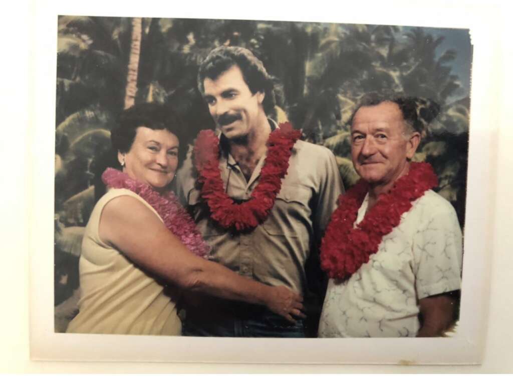 A man with a mustache stands between an elderly woman and an elderly man. The trio is wearing leis and smiling, with a tropical backdrop featuring palm trees. The elderly woman and man stand on either side of the younger man, embracing him warmly.