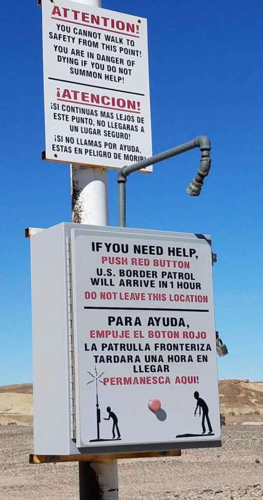 Signs in English and Spanish warn that it's unsafe to walk further, and offer help by pushing a red button. The U.S. Border Patrol will arrive within an hour. A button with a protective cover is attached below the English text. Desert landscape in the background.