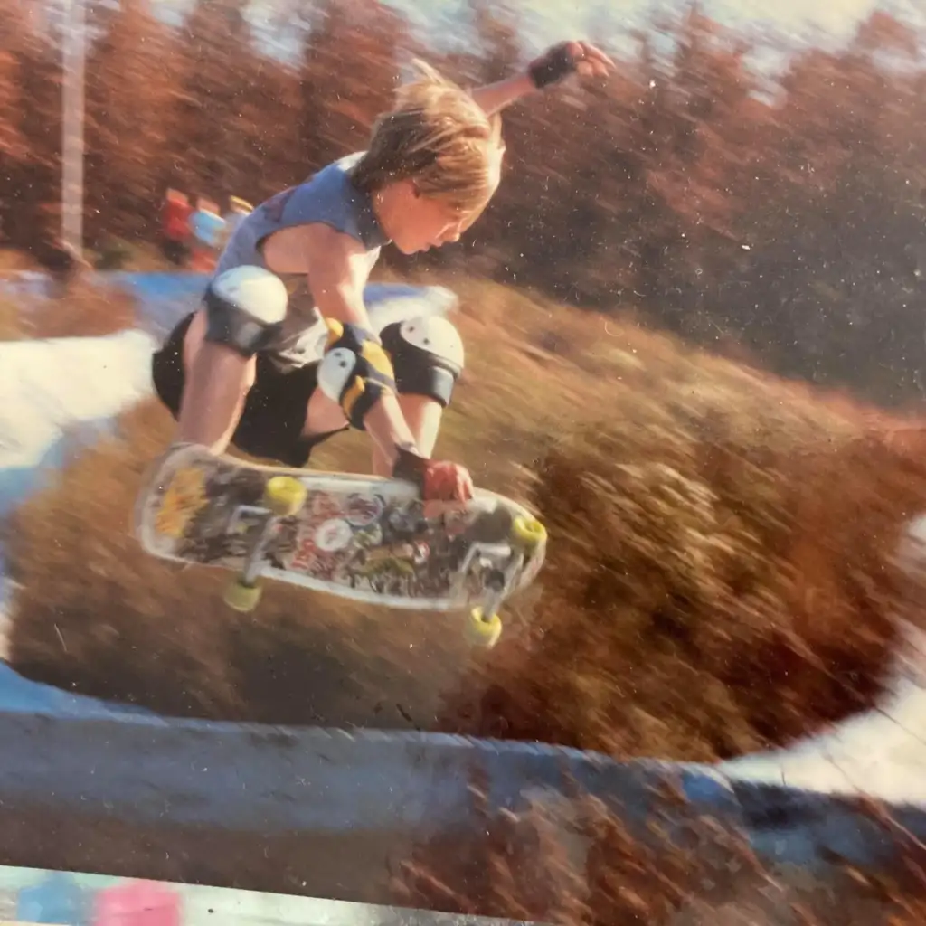 A young person with blonde hair performs an aerial trick on a skateboard in an outdoor skate park. They are wearing protective gear, including a helmet and knee pads. The background is blurred, featuring autumn-colored trees.