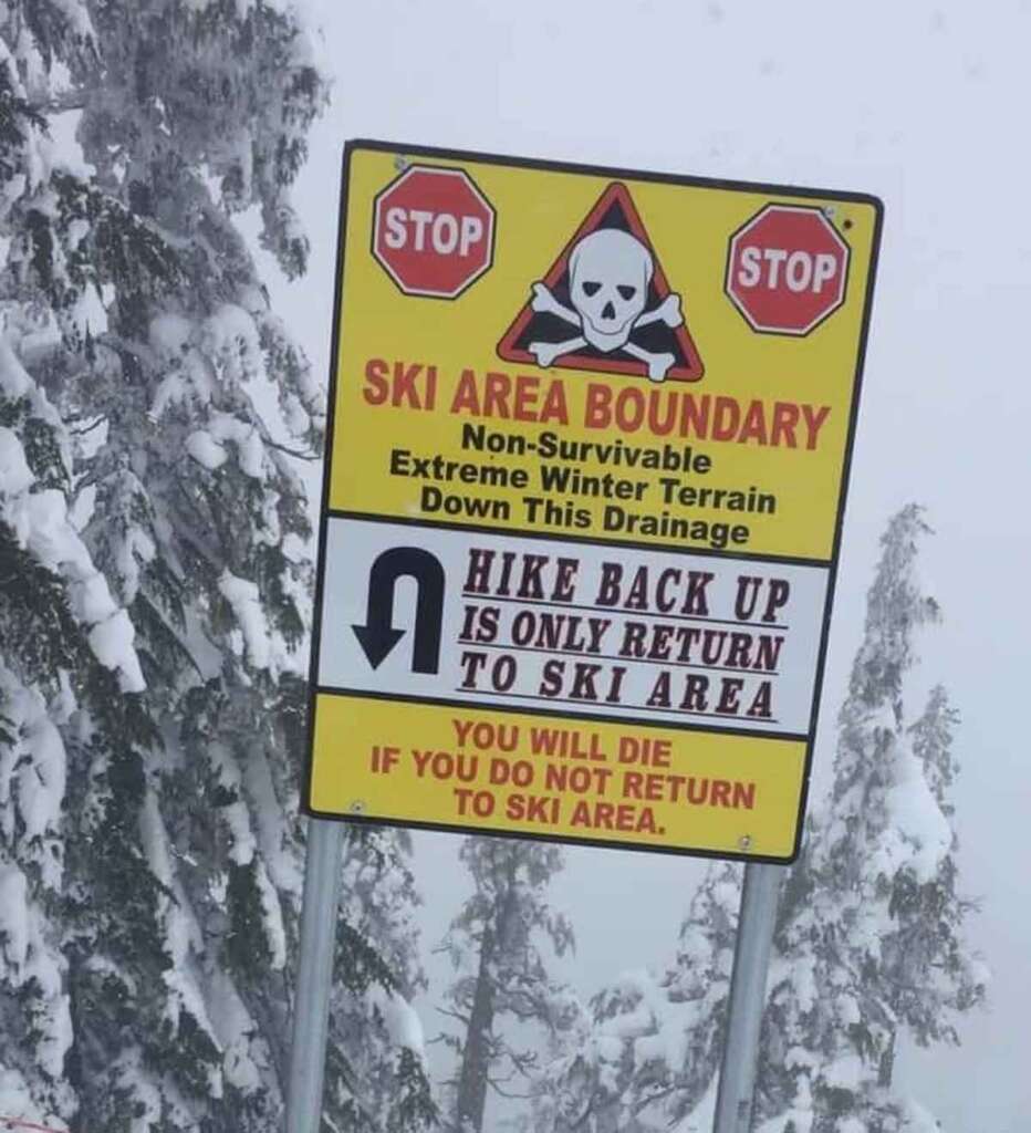 A warning sign in a snowy ski area bordered by trees. The top of the sign has two stop signs and a skull and crossbones symbol. Below it reads: "SKI AREA BOUNDARY. Non-Survivable Extreme Winter Terrain Down This Drainage. HIKE BACK UP IS ONLY RETURN TO SKI AREA. YOU WILL DIE IF YOU DO NOT RETURN TO SKI AREA.