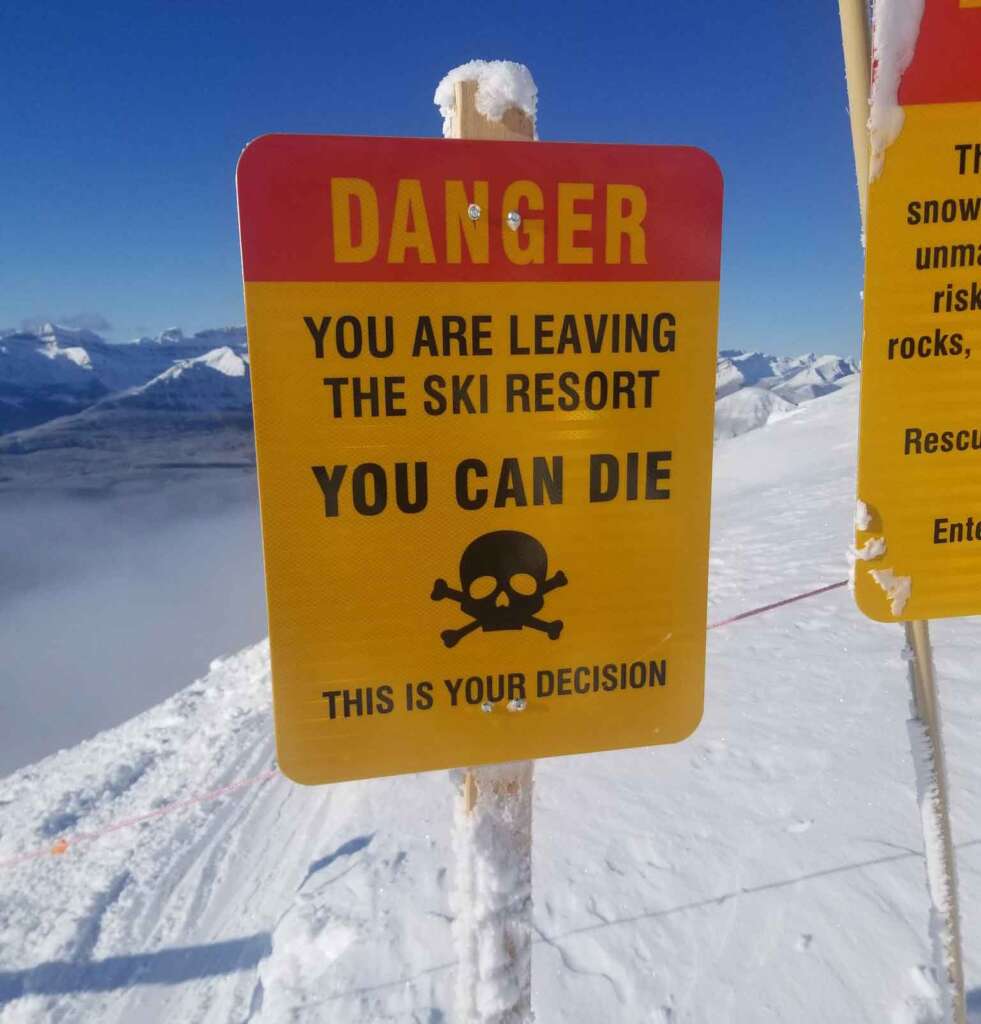 A yellow and red sign on a snowy mountain warns, "DANGER: YOU ARE LEAVING THE SKI RESORT. YOU CAN DIE. THIS IS YOUR DECISION," with a skull and crossbones symbol. Snow-covered peaks and a clear sky are visible in the background.