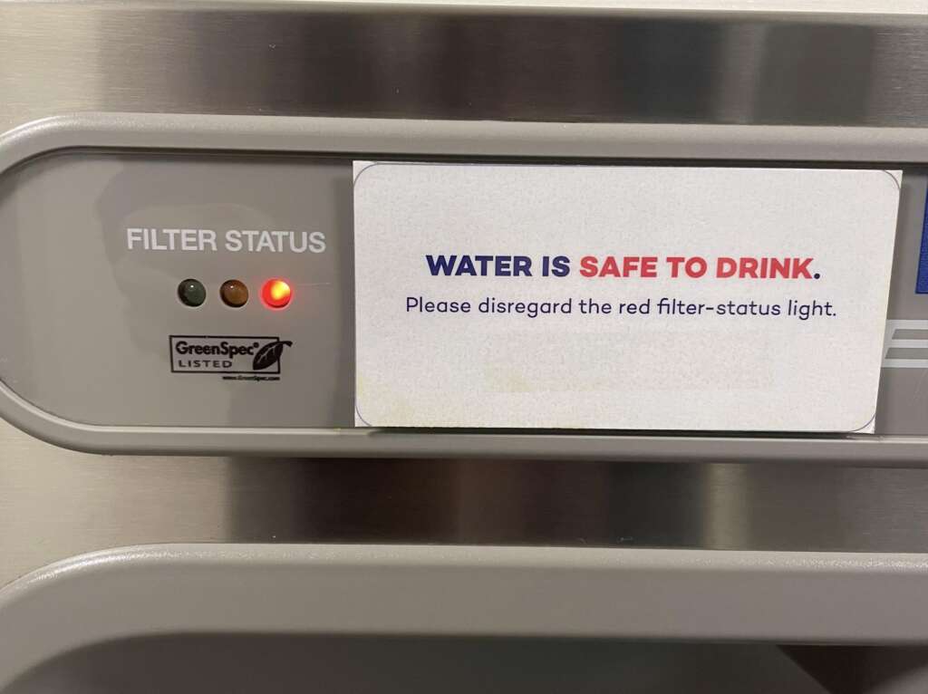Close-up of a filter status indicator on a panel. The indicator shows a red light, but there's a sign next to it that reads, "WATER IS SAFE TO DRINK. Please disregard the red filter-status light." A GreenSpec Listed label is also visible.