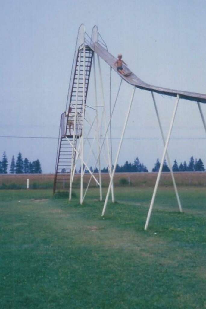 Two children are on a towering metal slide in a grassy park. One child is climbing up the steep ladder, while the other is already halfway down the slide. Trees and an open field are visible in the background.