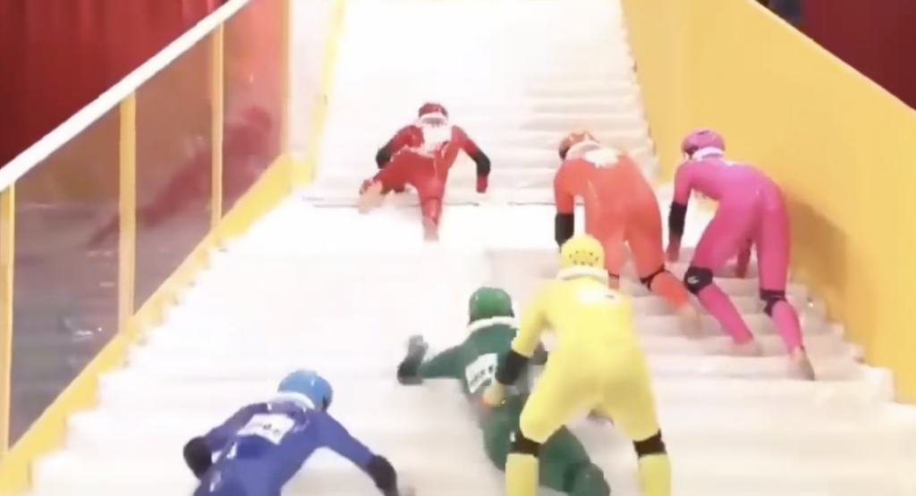 Five people in colorful outfits (blue, green, pink, yellow, and red) and helmets are climbing a set of stairs. Some are struggling, with one person in red almost at the top and others either on the stairs or at the bottom.