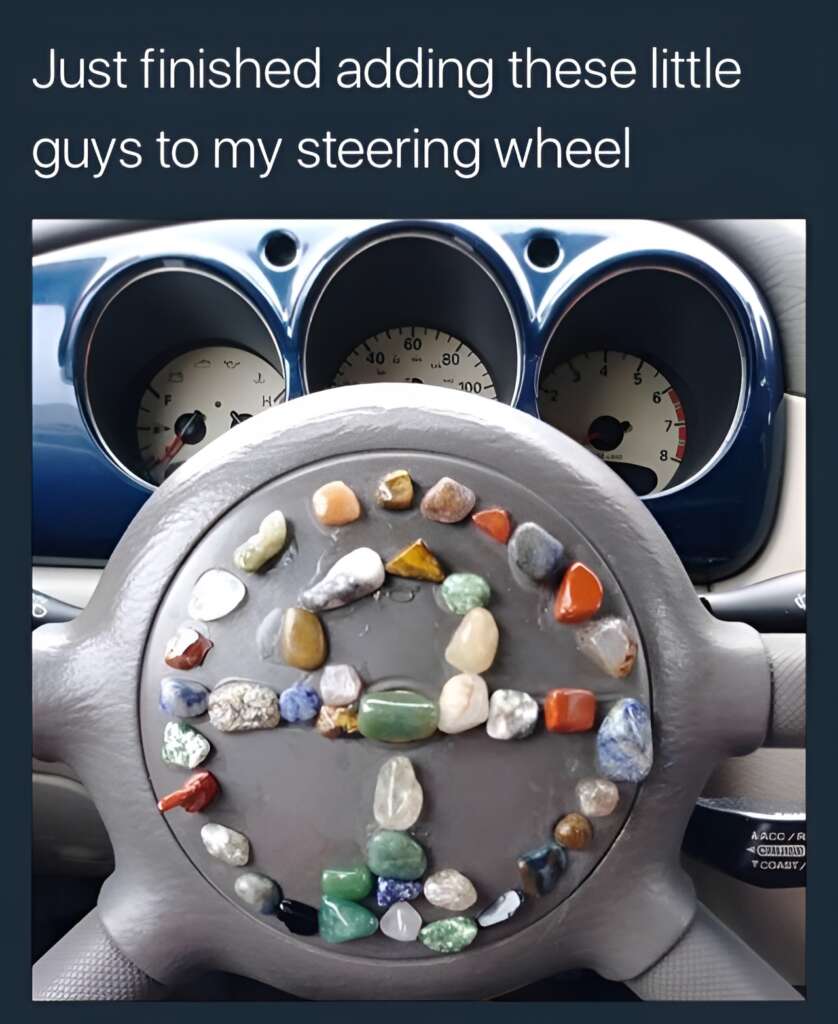 The image shows a car steering wheel decorated with various colorful stones and pebbles arranged in a pattern. The photo is taken from the driver's perspective, showing the dashboard and gauges behind the steering wheel. A caption reads, "Just finished adding these little guys to my steering wheel.