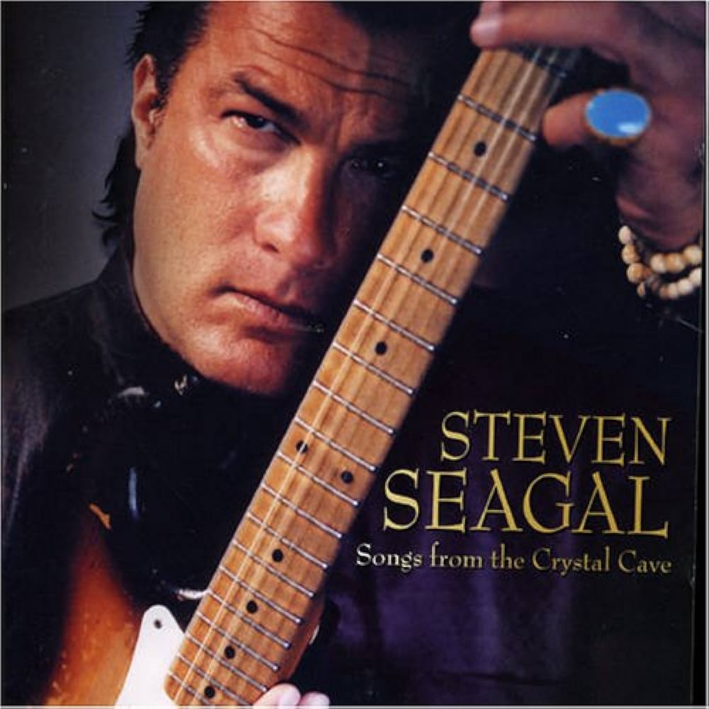 A man with slicked-back hair sternly looks at the camera while holding an electric guitar. The text on the image reads "Steven Seagal: Songs from the Crystal Cave." He is wearing a black shirt and a bracelet, with a light purple background.