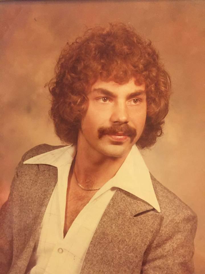 A man with curly hair and a mustache is posing for a portrait. He is wearing a grey blazer over a white shirt with a wide, pointed collar. The background is a warm, mottled brown.