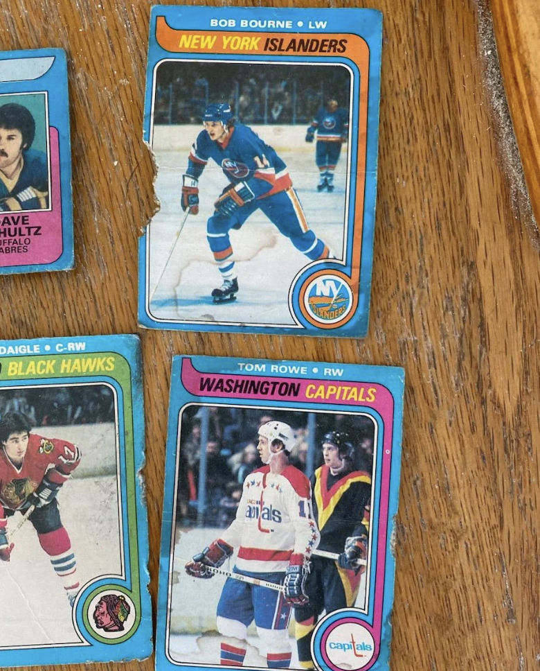 A close-up of three vintage hockey trading cards on a wooden surface. The top card features Bob Bourne of the New York Islanders. Beneath it, a card of Tom Rowe from the Washington Capitals. To the left, partially shown, is another card featuring a player from the Chicago Blackhawks.