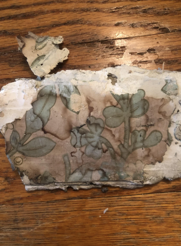 A close-up of a weathered, broken piece of wall plaster with a faded floral pattern. The piece rests on a wooden surface, with one smaller fragment lying separately above it. The floral design appears delicate and timeworn.