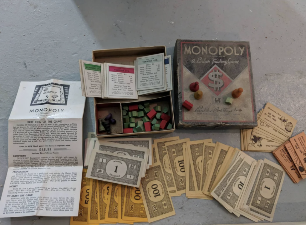 A vintage Monopoly game set is displayed, featuring a worn game box, various game pieces, property cards, Chance and Community Chest cards, paper money in different denominations, wooden houses and hotels, and the instruction manual.