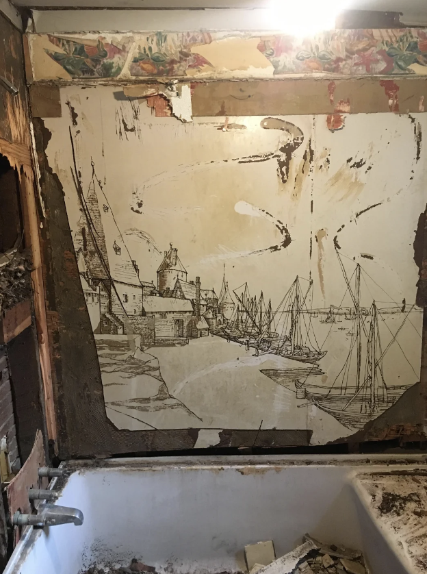 An old, partially demolished bathroom with a visible mural of a coastal town with boats on the water. The mural, possibly damaged during the demolition, features buildings, boats, and water. Debris and dirt are scattered around the bathtub and floor.