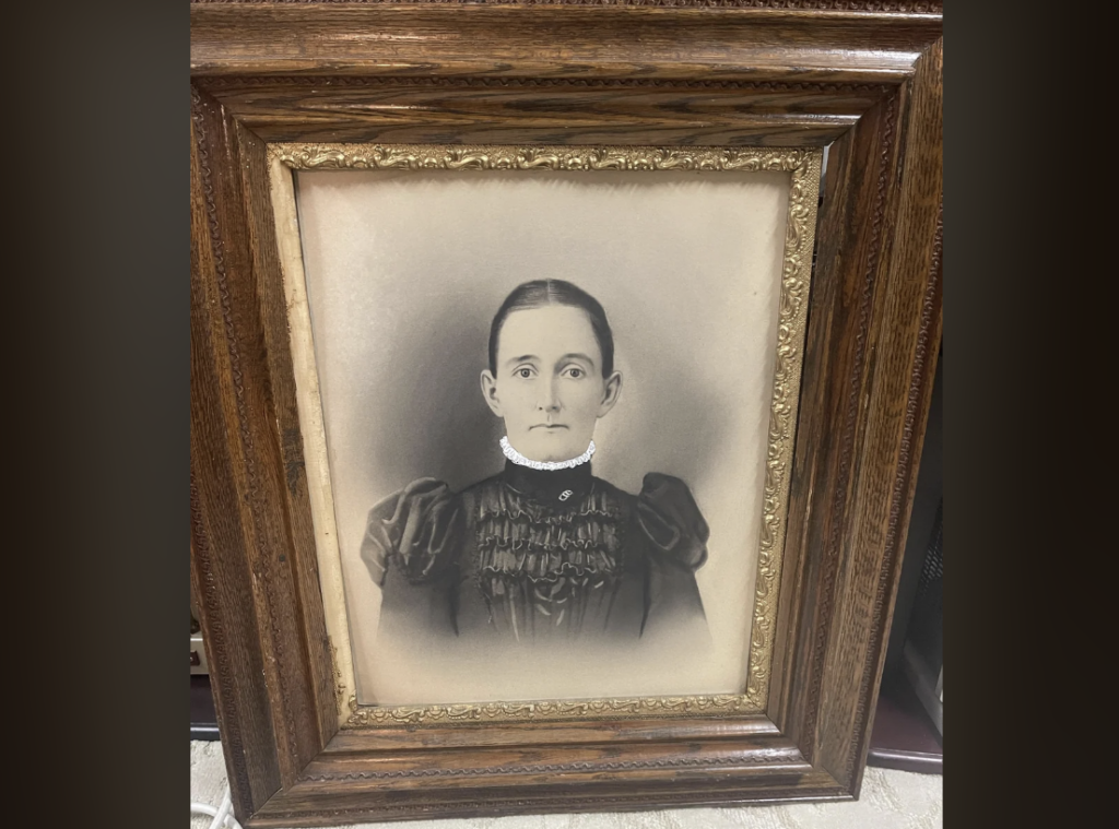 A sepia-toned portrait of a woman from the late 19th or early 20th century. She is wearing a high-necked dress with puffed sleeves and a lace collar. Her hair is pulled back tightly. The portrait is displayed in an ornate, dark wood frame with gilded trim.