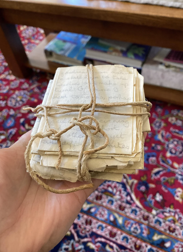 A hand holds a bundle of old, handwritten letters tied together with brown string. The background features a red, patterned carpet and the lower part of a wooden table with books stacked on a shelf underneath.