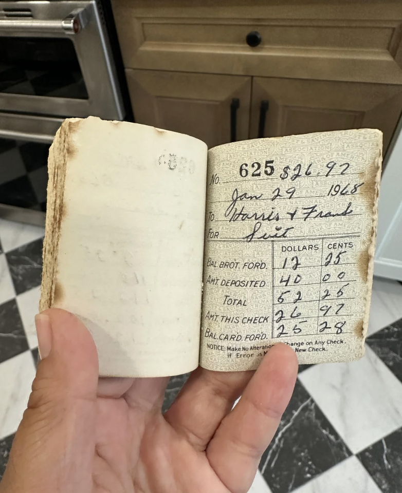 A hand is holding an old, worn checkbook open to a page with handwritten financial entries. The page lists transaction details such as dates, amounts deposited, and check numbers from 1968. The checkbook is held in front of a kitchen setting with a tiled floor and cabinetry.