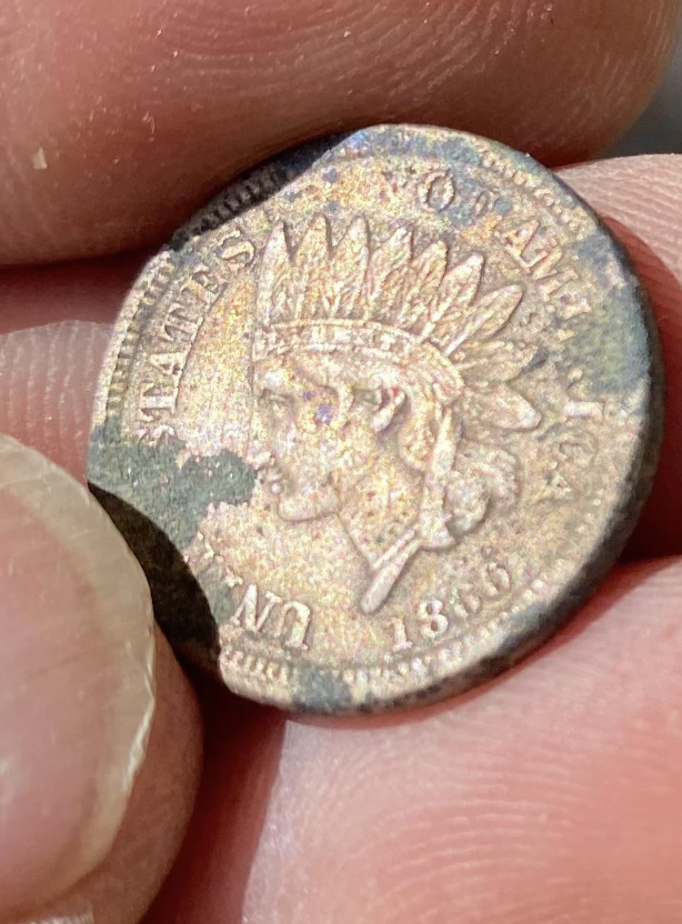 Close-up image of a hand holding a worn 1863 U.S. Indian Head penny. The coin features the profile of Lady Liberty wearing a Native American headdress and exhibits significant wear and tear, particularly around the edges, with a noticeable chunk missing.