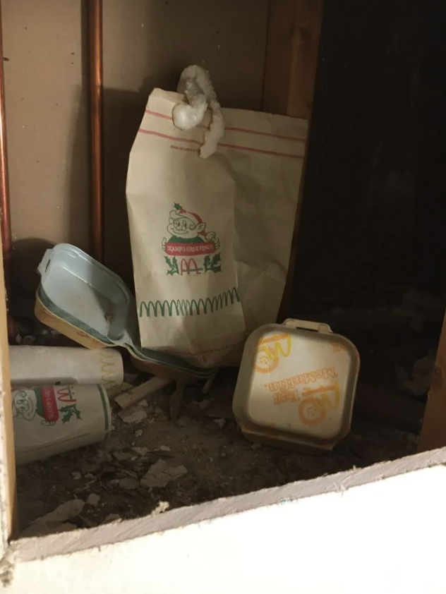 A hidden compartment in a wall reveals old McDonald's packaging, including cups, bags, and food containers amidst dust and debris. The items appear to be vintage, dating back to a previous era of the company's branding.