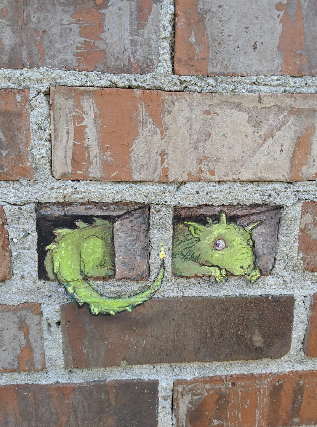 An image shows a small, green dragon sculpture peeking out from two adjacent bricks in a brick wall. The dragon's head and part of its body emerge from the gaps, giving the illusion that it is slithering out of the wall.